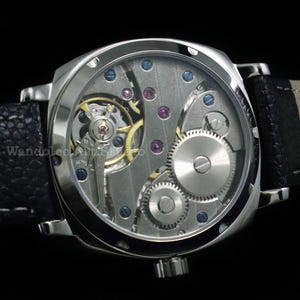Puede incluir: Un reloj de pulsera plateado con una correa de cuero negro. La esfera del reloj es transparente, revelando el intrincado movimiento mec&aacute;nico con engranajes, joyas y tornillos. La caja del reloj es cuadrada.