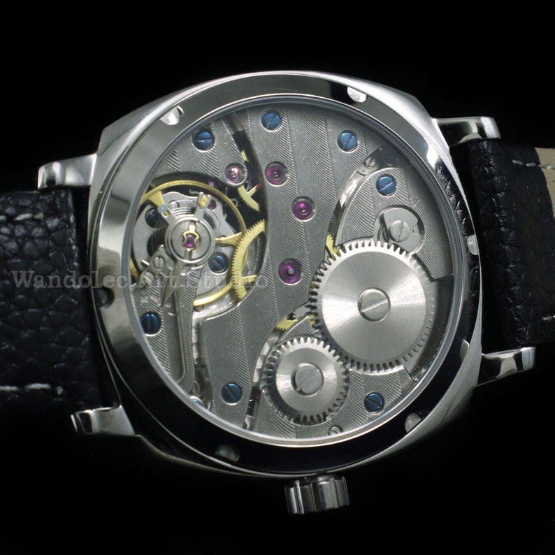 Puede incluir: Un reloj de pulsera plateado con una correa de cuero negro. La esfera del reloj es transparente, revelando el intrincado movimiento mec&aacute;nico con engranajes, joyas y tornillos. La caja del reloj es cuadrada.