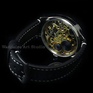 May include: A silver-toned wristwatch with a black leather band and white stitching. The watch face is open, revealing the intricate gold-colored gears and mechanisms inside. The text "Wandoleo Art Studio" is visible.