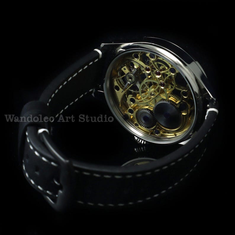May include: A silver-toned wristwatch with a black leather band and white stitching. The watch face is open, revealing the intricate gold-colored gears and mechanisms inside. The text "Wandoleo Art Studio" is visible.