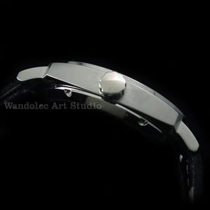 Puede incluir: Vista lateral de un reloj de pulsera plateado con una correa de cuero negro. El reloj tiene una esfera rectangular y una corona redonda. El texto "Wandolec Art Studio" est&aacute; impreso en la esfera.