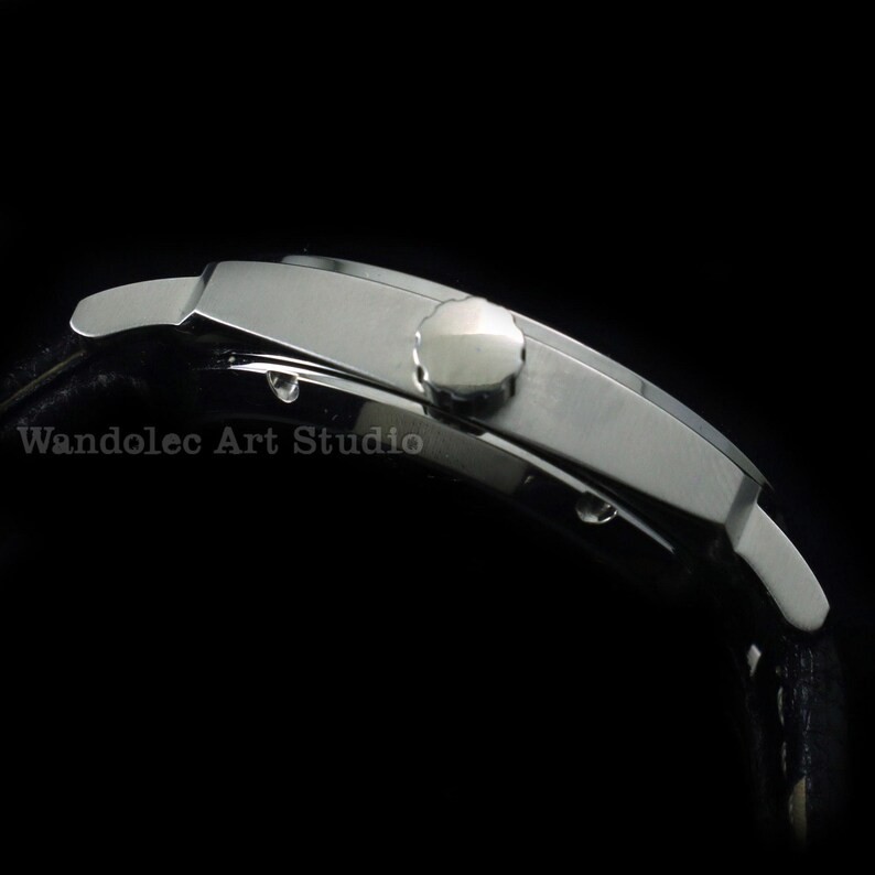 Puede incluir: Vista lateral de un reloj de pulsera plateado con una correa de cuero negro. El reloj tiene una esfera rectangular y una corona redonda. El texto "Wandolec Art Studio" est&aacute; impreso en la esfera.