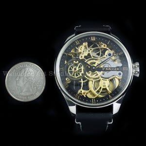 May include: A skeleton watch with a black face and gold-colored gears, featuring Roman numeral hour markers. The watch has a silver-colored case and a black leather band. A quarter is next to the watch for size comparison.