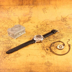 Puede incluir: Un reloj plateado con una correa de cuero negro, una peque&ntilde;a caja decorativa y un colgante de br&uacute;jula en una cadena descansan sobre un mapa antiguo. La esfera del reloj revela el mecanismo interno. La caja tiene detalles ornamentales.