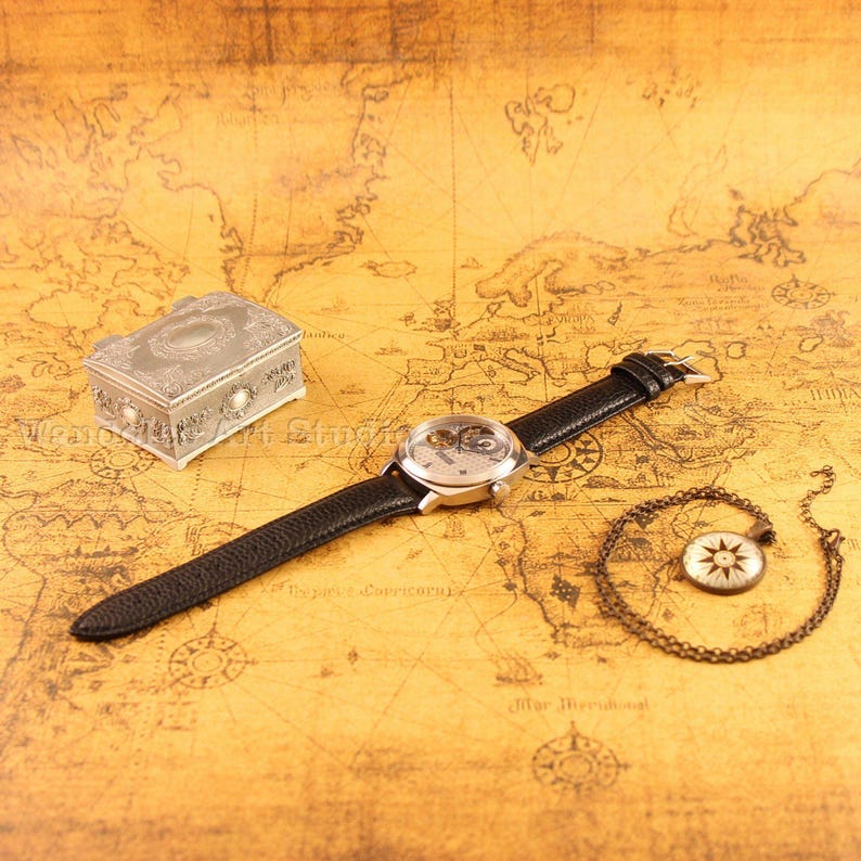 Puede incluir: Un reloj plateado con una correa de cuero negro, una peque&ntilde;a caja decorativa y un colgante de br&uacute;jula en una cadena descansan sobre un mapa antiguo. La esfera del reloj revela el mecanismo interno. La caja tiene detalles ornamentales.