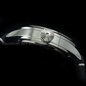 May include: Close-up of a silver wristwatch with a black leather band. The watch features a textured crown and a curved, polished case. The band is black with white stitching. The watch is set against a black background.
