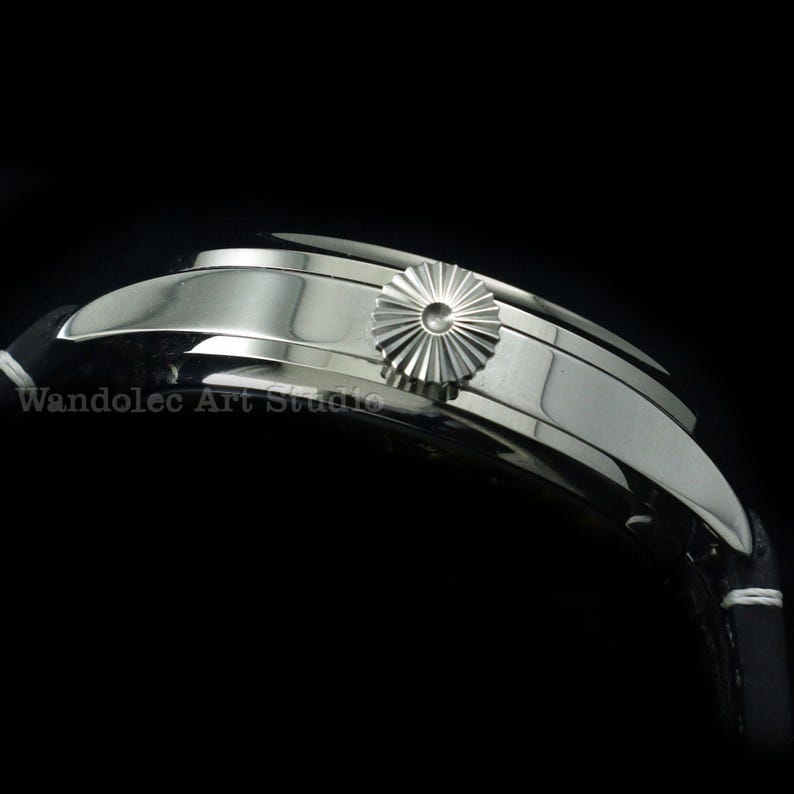 May include: Close-up of a silver wristwatch with a black leather band. The watch features a textured crown and a curved, polished case. The band is black with white stitching. The watch is set against a black background.