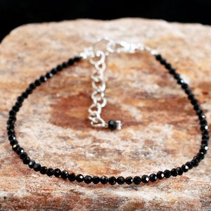 Op de afbeelding: Een verfijnde armband bestaande uit een enkele streng kleine, gefacetteerde zwarte kralen. De armband is voorzien van een zilverkleurige metalen karabijnsluiting en een korte verlengketting met grotere zilverkleurige schakels voor verstelbaarheid.