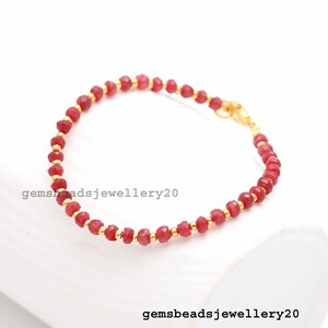 Puede incluir: Un brazalete de cuentas de piedras preciosas rojas con detalles dorados. El brazalete está hecho con cuentas pequeñas y facetadas y tiene un cierre de langosta.