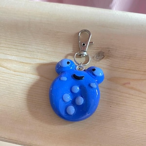 May include: A blue, round keychain with a cartoon-like design. The keychain features two small, round protrusions on top and several smaller circles on the front. A silver-colored clasp is attached to the top.