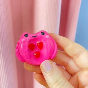 May include: A glossy, pink frog-shaped figurine with three red heart accents. The frog has black eyes and small pink cheeks. The figurine is held in a hand against a pink background.
