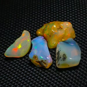 Könnte beinhalten: Vier rohe Opale in verschiedenen Formen und Farben werden vor einem dunklen, strukturierten Hintergrund gezeigt. Die Opale reichen von Weiß mit Farbblitzen bis hin zu Blau und Gelb und zeigen ihre natürliche Schönheit. Diese Edelsteine eignen sich ideal für die Schmuckherstellung.