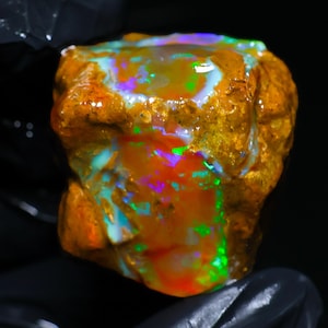 May include: A raw, unpolished boulder opal gemstone featuring a textured, golden-brown matrix with vibrant, iridescent flashes of neon green, electric blue, and bright red play-of-colour across its irregular, multifaceted surface.