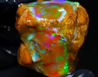 May include: A raw, unpolished boulder opal gemstone featuring a textured, golden-brown matrix with vibrant, iridescent flashes of neon green, electric blue, and bright red play-of-color across its irregular, multifaceted surface.
