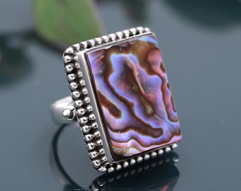 Könnte beinhalten: Ein silberner Ring mit einem rechteckigen Einlegeteil aus Abalone-Muschel. Die Muschel hat schillernde Farben in Lila, Braun und Blau. Der Ring hat einen Perlenrand.