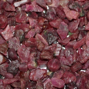 May include: A close-up view of a pile of small, rough, red ruby gemstones. The rubies are various shapes and sizes, with some having a slightly darker, purplish hue.