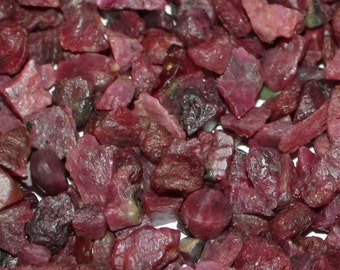 May include: A close-up view of a pile of small, rough, red ruby gemstones. The rubies are various shapes and sizes, with some having a slightly darker, purplish hue.
