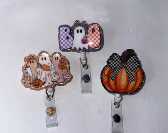 May include: Three Halloween-themed badge reels. One features ghosts and pumpkins, another says "Boo" with a ghost, and the third is a pumpkin with a checkered bow. Each reel has a clear plastic clip.