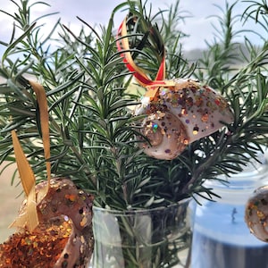 May include: Decorative seashell ornaments with glitter accents hang from rosemary sprigs in a glass vase. The ornaments have gold, brown, and iridescent glitter. The rosemary is green and fresh. The ornaments are suspended by ribbons.