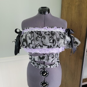 May include: A two-piece top and sash set displayed on a mannequin. The top is off-the-shoulder with black ribbon ties, a black and gray swirl pattern, and lavender lace trim. The sash mirrors the top's design.