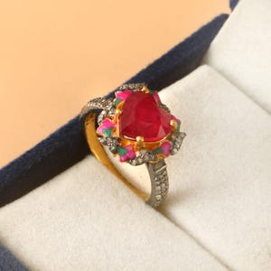Puede incluir: Anillo con gema de rubí en forma de corazón, con banda dorada y engaste plateado. El rubí está rodeado por un diseño floral con detalles rosas, verdes y blancos. El anillo se presenta en una caja blanca con interior azul.