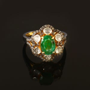 May include: A gold and silver ring featuring a large oval-cut emerald center stone. The emerald is surrounded by smaller, round, clear gemstones and a halo of smaller stones. The ring is set against a black background.