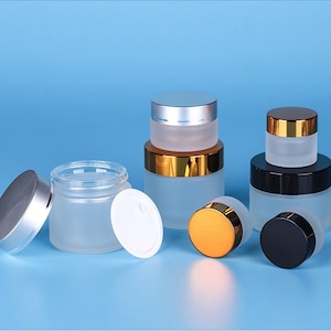May include: Several frosted glass jars with silver, gold, and black lids are displayed against a blue background. The jars vary in size and shape, with some lids off and some on. The jars are designed for cosmetics or skincare products.