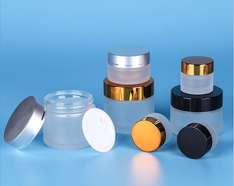 May include: Several frosted glass jars with silver, gold, and black lids are displayed against a blue background. The jars vary in size and shape, with some lids off and some on. The jars are designed for cosmetics or skincare products.