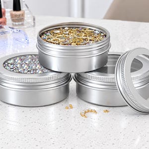 May include: Three round silver metal tins with clear windows on the lids. One tin is filled with gold-toned charms and star shapes, another with iridescent rhinestones, and the third is empty with its lid placed beside it on the surface.