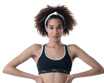 May include: A woman with curly brown hair is wearing a black sports bra with white trim and the text "Wendy" on the band. She is also wearing a white headband tied in a knot on top of her head.