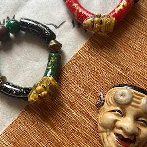 Op de afbeelding: Drie decoratieve armbanden en een masker. Eén armband is rood met gouden accenten en een gebeeldhouwd gezicht. Een andere is zwart met gouden spikkels en een gebeeldhouwd gezicht. De derde armband is groen met gouden accenten. Er is ook een lachend masker aanwezig.