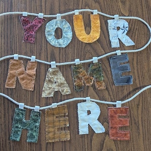 Puede incluir: Una pancarta de tela con el texto YOUR NAME HERE deletreado en letras individuales con diversos patrones y colores. Cada letra está unida a una cuerda blanca gruesa mediante pequeñas presillas de tela, mostrando texturas y estampados.