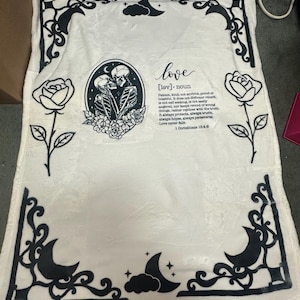 Op de afbeelding: Witte deken met zwarte decoratieve rand, rozen, manen en wolken. Het midden toont een zwart-wit illustratie van twee omarmende skeletten, omringd door bloemen, met het woord "love" en definitietekst.