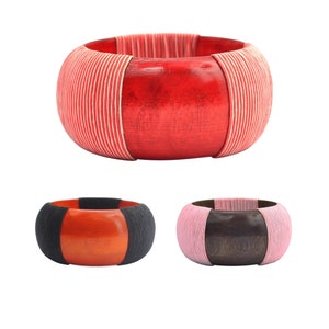Könnte beinhalten: Drei Holz-Armbänder mit verschiedenen Farbstreifen. Das erste Armband ist rot mit weißen Streifen, das zweite ist orange mit schwarzen Streifen und das dritte ist braun mit rosa Streifen.