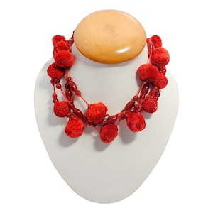 May include: A red necklace with multiple strands of small red beads and larger, textured red pom-poms. The necklace is displayed on a white jewelry bust, with a large, round, light-colored wooden pendant at the top.
