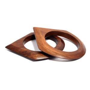 Może przedstawiać: Dwa drewniane bransoletki, jedna na drugiej, obie w kształcie diamentu z zaokrąglonymi krawędziami. Bransoletki są w kolorze ciemnego brązu.