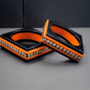 May include: Two square bangles. The bracelets are black with an orange interior and a row of small, silver-coloured beads. The bangles are displayed on a grey surface with a dark background.