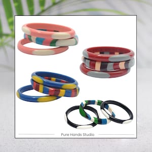 Peut inclure: Ensemble de quatre bracelets rigides colorés avec des motifs géométriques. Les bracelets sont en plastique et ont une finition brillante. Les bracelets sont empilés les uns sur les autres, le bracelet du haut étant rose avec des pois blancs, le deuxième bracelet étant blanc avec des rayures bleues et vertes, le troisième bracelet étant bleu avec des rayures jaunes et rouges, et le quatrième bracelet étant noir avec des pois blancs. Les bracelets sont disposés en forme de carré, avec le texte "Pure Hands Studio" écrit en dessous des bracelets.