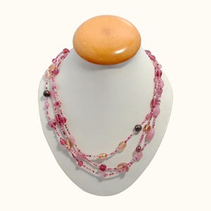 May include: A multi-strand necklace with pink and purple beads of various shapes and sizes. The necklace is displayed on a white bust with a wooden top. The beads range in color from light pink to deep purple.