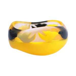 Puede incluir: Un brazalete de plástico amarillo, negro y blanco con un diseño de pájaro.