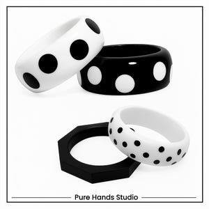 May include: A collection of black and white bangles with a polka dot design. The image features a white bangle with black dots, a black bangle with white dots, and a white bangle with black dots. A black, geometric, hexagonal ring is also present. The text "Pure Hands Studio" is at the bottom.