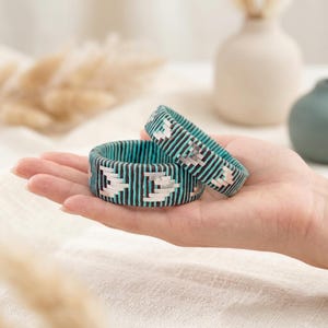 Puede incluir: Dos pulseras tejidas turquesa y negras con un patrón geométrico. Las pulseras están apiladas una encima de la otra, mostrando el intrincado diseño. Las pulseras miden aproximadamente 2.5 cm de ancho.