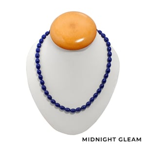 May include: A navy blue beaded necklace displayed on a white bust. The necklace features oval-shaped beads. A large, round, light brown wooden element is at the top of the bust. The text "MIDNIGHT GLEAM" is at the bottom.