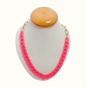 Pode incluir: Um colar de corrente rosa brilhante exibido em um manequim branco. A corrente grossa é feita de elos interligados e é conectada a uma corrente dourada em cada extremidade. O manequim tem um topo de madeira.