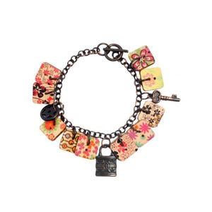 Puede incluir: Un brazalete de cadena de cobre con múltiples dijes. Los dijes son cuadrados y presentan un patrón floral en tonos de rosa, amarillo y verde. También hay un pequeño dije de candado y un dije de llave.