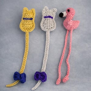 May include: Three handmade crochet bookmarks. Two are yellow and light gray cat-shaped figures with purple bows at the base. The third is a pink flamingo with a black and white beak and two long dangling legs. Each piece is crafted with yarn.