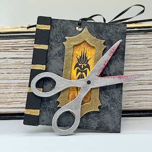May include: A black book with gold accents and a pair of silver scissors with a red blade. The book has a gold frame with a yellow and orange design inside.