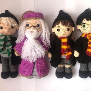 May include: Four crocheted plush toys inspired by characters from the Harry Potter series. The toys are all wearing black robes and scarves in different colors. The characters include a wizard with a white beard, a young witch with brown hair, and two young wizards with black hair.
