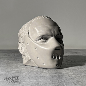 May include: A white ceramic sculpture of a person's head wearing a mask with a metal grill over the mouth. The mask has holes around the eyes and mouth.