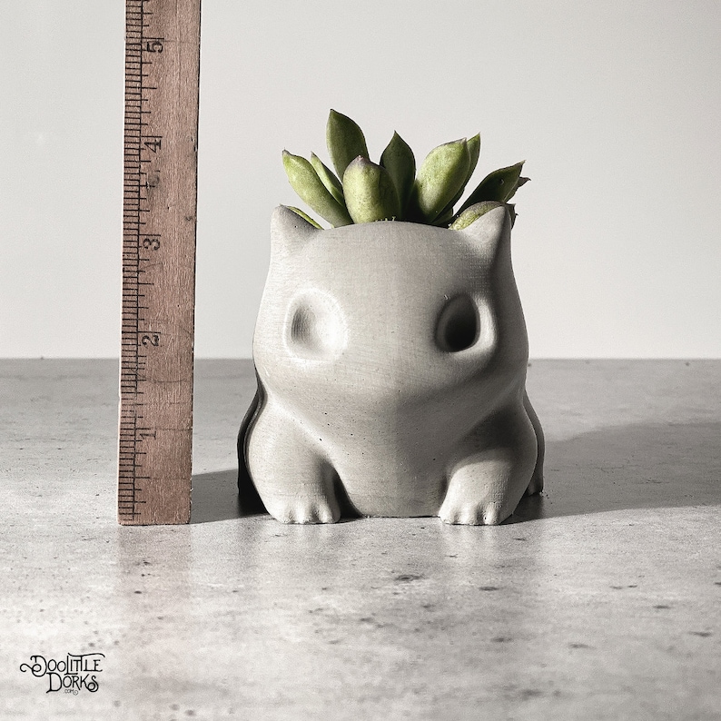 Puede incluir: Una maceta de hormig&oacute;n gris con forma de personaje de dibujos animados, que contiene una planta suculenta verde. La maceta mide aproximadamente 10 cm de altura. El logotipo "DooLittle Dorks" es visible en la parte inferior izquierda.